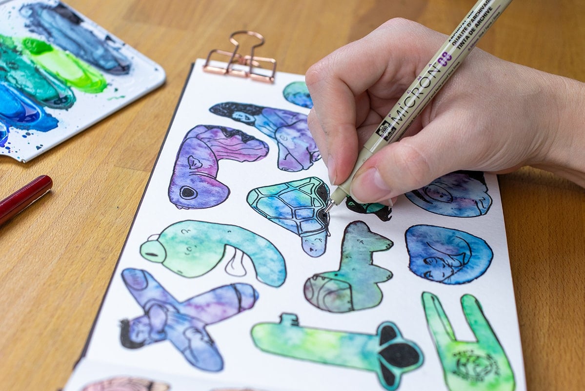 Hand holding pen drawing tortoise out of watercolor blob.